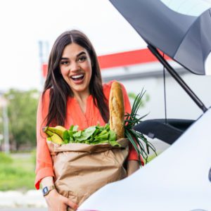 Uma mulher sorridente est&aacute; parada ao lado do porta-malas aberto de um carro, segurando uma sacola de papel cheia de mantimentos, incluindo uma baguete, bananas e folhas verdes.