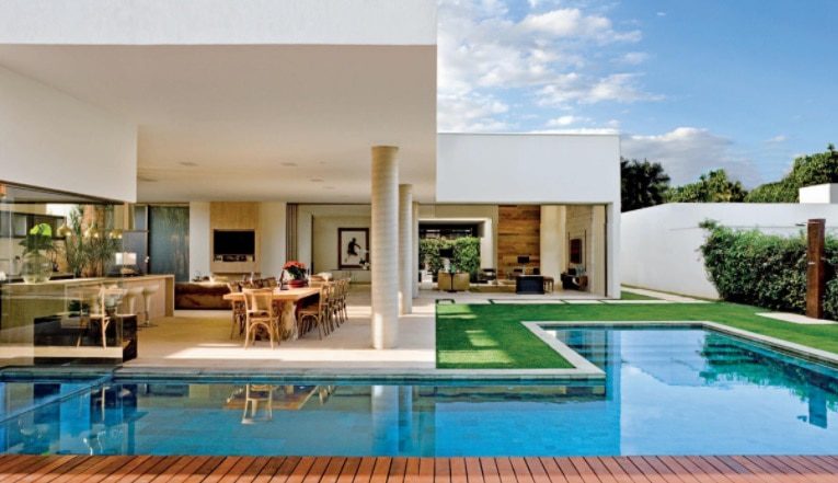 Casa moderna com design aberto, com grandes paredes de vidro, área de jantar ao ar livre, gramado verde, deck de madeira e uma piscina retangular — verdadeiramente uma das Casas dos sonhos de Brasília — situada sob um céu azul com algumas nuvens.