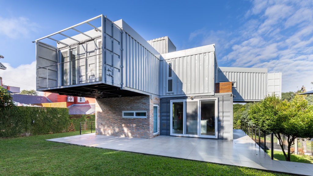 Uma casa moderna, construída com contêineres e tijolos empilhados, ergue-se sobre um gramado, com grandes janelas, um terraço e um pavimento superior parcialmente suspenso — um verdadeiro exemplo das Casas dos Sonhos de Porto Alegre. O céu está parcialmente nublado.