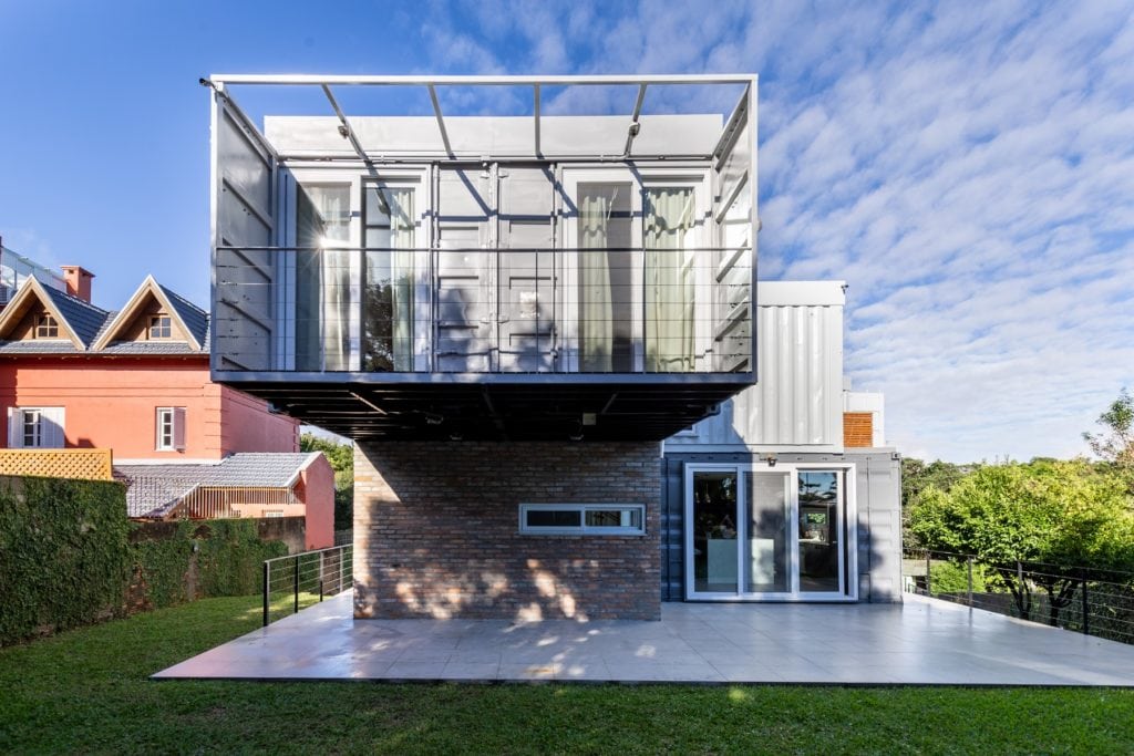 Casa moderna de dois andares com um grande andar superior de vidro e metal que se estende sobre um andar térreo de tijolos e vidro, cercada por vegetação em um dia ensolarado — uma das casas dos sonhos de Porto Alegre.