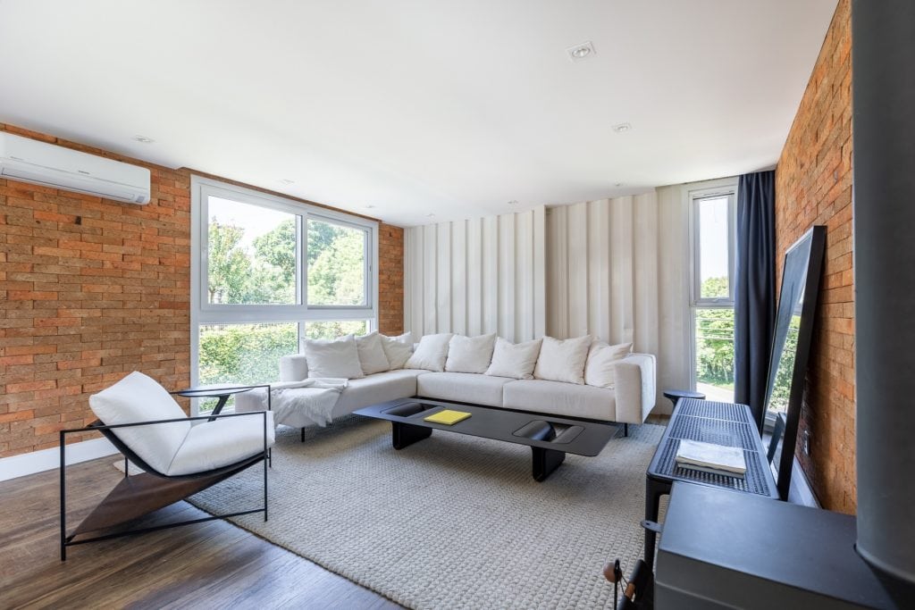 Uma sala de estar moderna com um grande sofá secional branco, mesa de centro preta, poltrona minimalista e grandes janelas que deixam entrar luz natural — inspiração perfeita para as Casas dos sonhos de Porto Alegre, com paredes de tijolos, piso de madeira e um tapete claro.