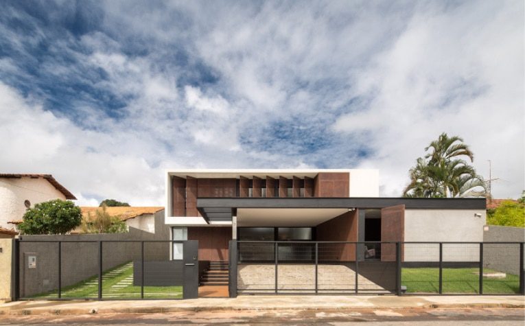 Casa moderna de dois andares com telhado plano, janelas grandes e uma mistura elegante de paredes externas brancas, marrons e cinzas — uma das casas dos sonhos de Brasília — atrás de uma cerca de metal, com uma entrada de automóveis e pequenos gramados verdes em ambos os lados.