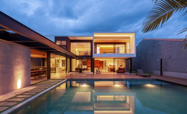 Casa moderna de dois andares com grandes janelas de vidro e iluminação interna aconchegante, com vista para uma piscina ao entardecer. Mesas ao ar livre e palmeiras evocam as Casas dos Sonhos de Brasília, criando uma atmosfera luxuosa e convidativa.