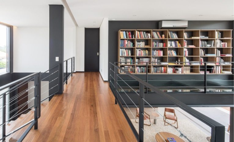Um corredor moderno e aberto com piso de madeira e grades pretas, este espaço reflete a elegância das Casas dos sonhos de Brasília, com uma grande estante e uma área de leitura iluminada e arejada com cadeiras abaixo.