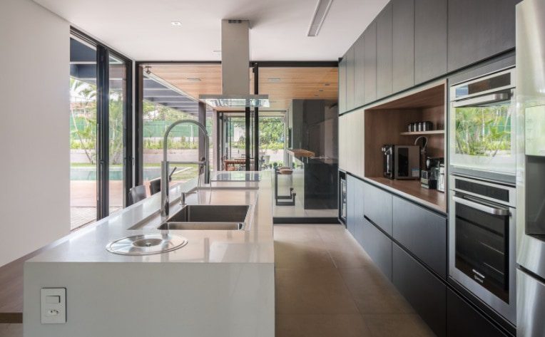 Cozinha moderna com armários escuros e elegantes, eletrodomésticos embutidos de aço inoxidável, uma espaçosa ilha branca com pia e fogão, janelas grandes e vista para um pátio e área verde — perfeita para quem busca as casas dos sonhos de Brasília.
