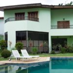 Uma moderna casa verde de dois andares com grandes janelas, varandas e persianas de madeira, cercada por arbustos paisagísticos e uma piscina com duas espreguiçadeiras brancas em um pátio de pedra em primeiro plano.