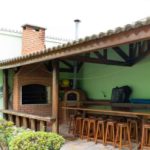 Pátio externo coberto com churrasqueira de tijolos e forno de pizza, balcão longo de madeira com vários bancos de bar, geladeira pequena e plantas de jardim em primeiro plano.