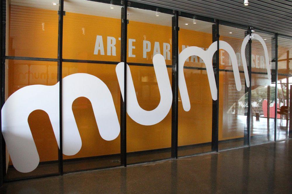 Grandes letras brancas com a inscrição "múmia" aparecem em vitrais com fundo laranja, possivelmente em um dos Bairros Nobres de Curitiba. Parte do texto "ARTE PAR" sugere um local ou exposição relacionada a artes.