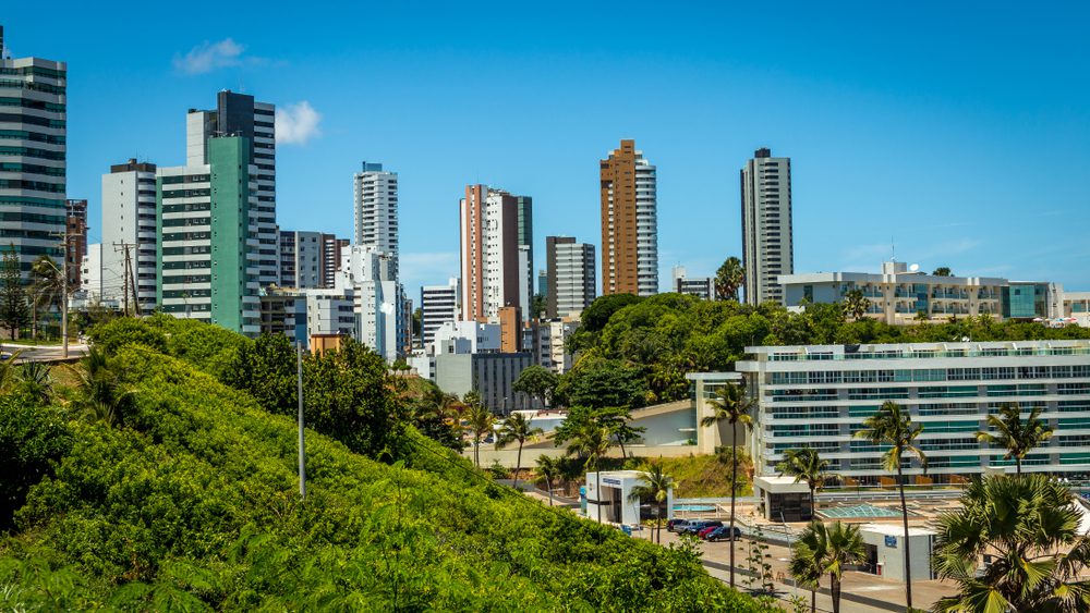 Uma paisagem urbana com arranha-céus modernos e hotéis atrás de árvores verdejantes e colinas sob um céu azul claro, com palmeiras e alguns carros em primeiro plano, captura o charme dos melhores bairros de Salvador.