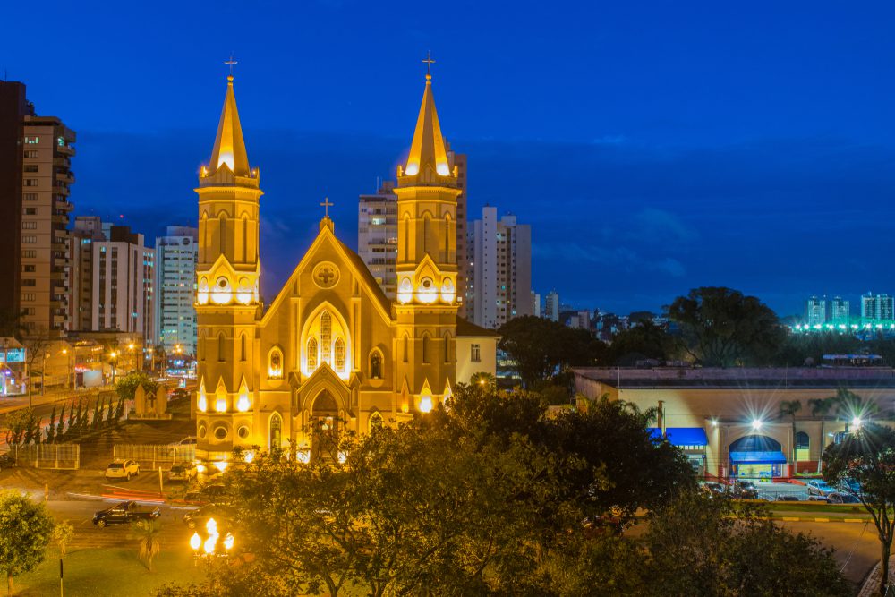 Uma grande igreja neogótica iluminada com torres gêmeas se destaca contra um céu azul profundo da noite, aninhada entre edifícios e árvores modernas da cidade em um dos bairros nobres de Curitiba.