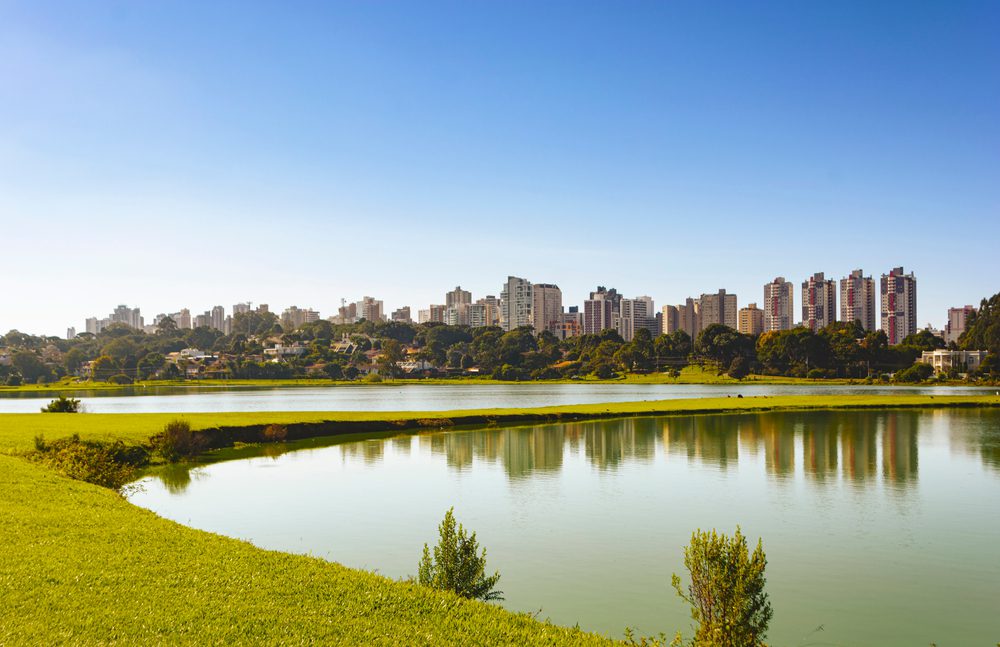 Um lago calmo com margens verdejantes e gramadas está em primeiro plano, refletindo o céu azul claro. Ao fundo, os bairros nobres de Curitiba formam um horizonte deslumbrante acima de uma fileira de árvores.