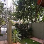 Pequeno jardim com plantas e árvores verdes, ao lado de um muro cinza e um portão de metal. Ao fundo, através da folhagem, é possível ver a calçada e os altos prédios de apartamentos.