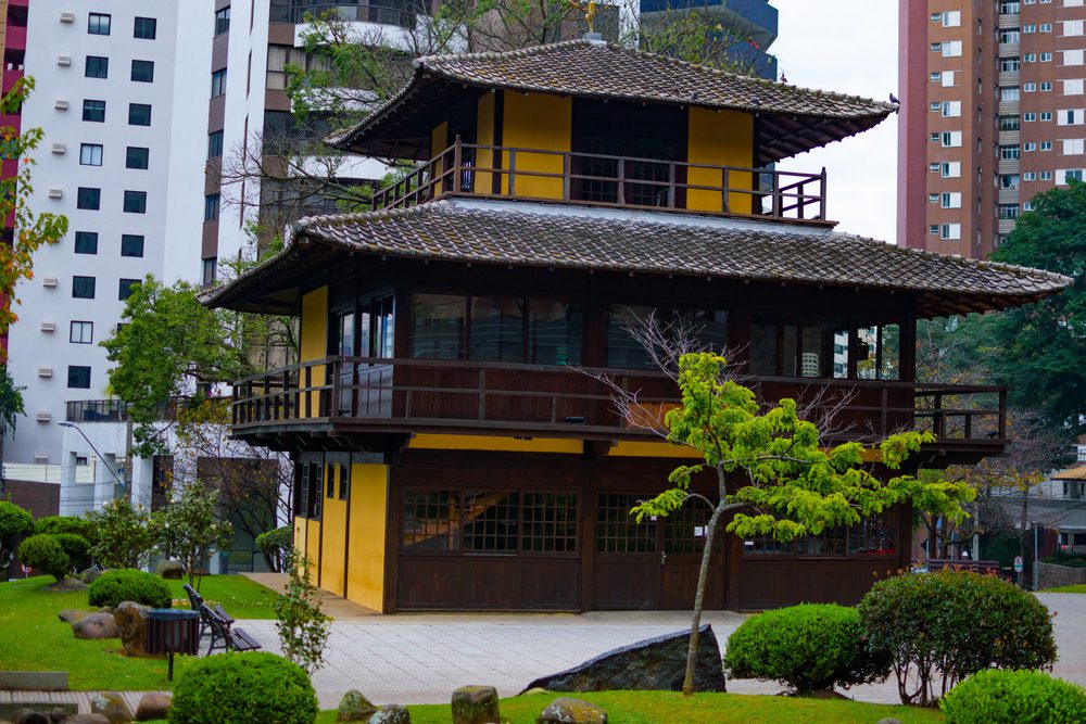 Um edifício tradicional em estilo japonês, com paredes amarelas e detalhes em madeira, fica em um jardim paisagístico em um dos bairros nobres de Curitiba, cercado por arranha-céus modernos.