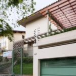 Uma casa de dois andares com porta de garagem verde, portão de entrada e terraço com pérgola de madeira. Placas na cerca indicam que a propriedade está disponível para venda ou aluguel. Árvores e plantas circundam a casa.