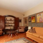 Uma sala de estar aconchegante com um sofá laranja, duas poltronas estampadas, uma mesa de centro redonda de madeira, quadros de parede e uma prateleira de madeira embutida aberta com itens decorativos. O piso é de madeira com um tapete estampado.