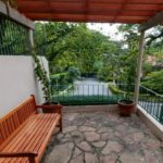 Um banco de madeira fica em um pátio de pedra sob uma pérgola de madeira, com plantas em vasos por perto, árvores verdes e uma entrada de automóveis visível ao fundo.