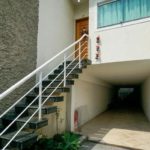 Uma escada externa com corrimão branco leva a uma porta de madeira no primeiro andar. Abaixo, há uma entrada coberta para carros ou garagem que se estende até os fundos, ao lado de uma parede texturizada e uma pequena planta verde.