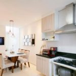 Cozinha e sala de jantar modernas com armários de madeira clara, bancada preta, fogão a gás, coifa de aço inoxidável, mesa de jantar com cadeiras bege, TV de parede e luminária pendente. Design claro, clean e minimalista.