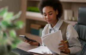 Imagem de uma mulher negra sentada em uma mesa, segurando uma calculadora e analisando documentos sobre alugar ou financiar, com um caderno aberto e um lápis à sua frente, parecendo concentrada.
