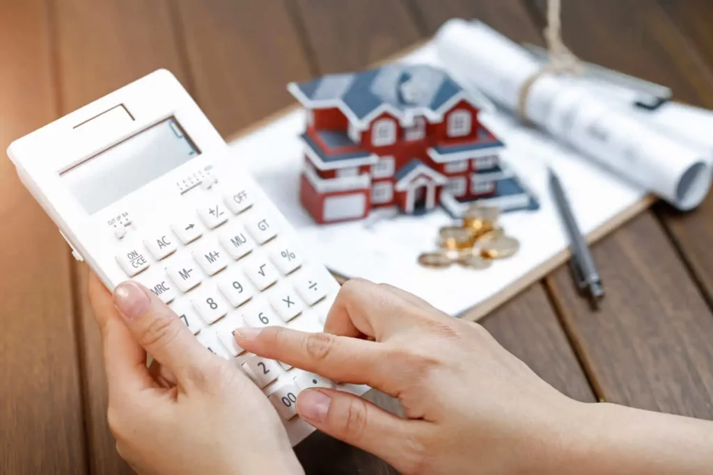 Imagem de uma pessoa usando uma calculadora em uma mesa com um modelo de casa pequena, moedas, caneta e documentos enrolados