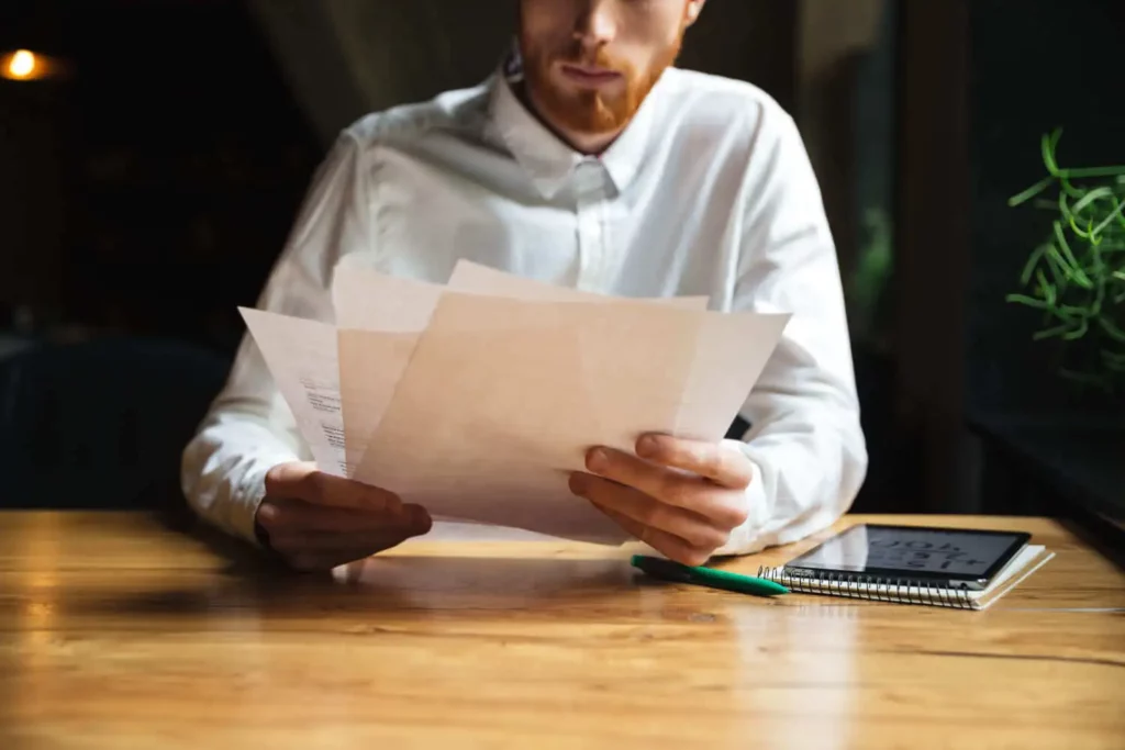 Imagem de um homem sentado enquanto analisa alguns papéis apoiado em cima de uma mesa de madeira para ilustrar matéria sobre certidão vintenária do imóvel
