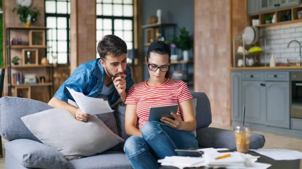 Imagem de um casal formado por um homem e uma mulher sentados no sofá da sala de casa analisando as contas em papéis e em um tablet para ilustrar matéria sobre como comprar um apartamento sem entrada
