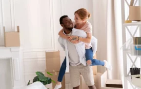 Imagem de um homem sorridente e uma mulher em um quarto com caixas de mudança e decoração branca para ilustrar matéria sobre imóvel de leilão. Eles parecem felizes e afetuosos.