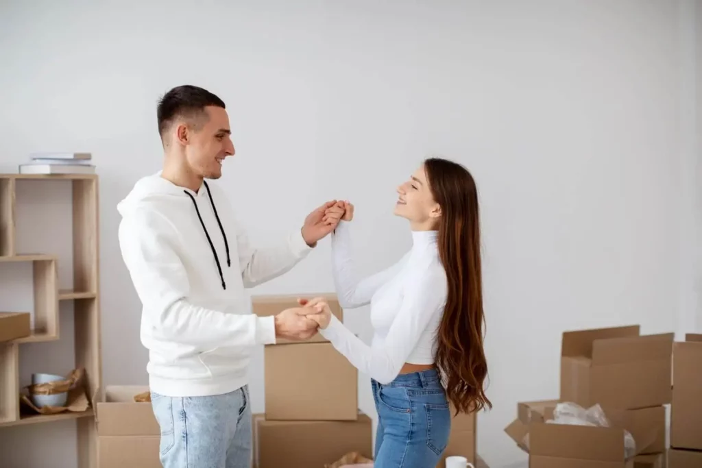 Imagem de um casal sorridente dá as mãos e dança em uma sala cercada por caixas de papelão embaladas para ilustrar matéria sobre imóvel de leilão da Caixa. Prateleiras com alguns itens são visíveis ao fundo.
