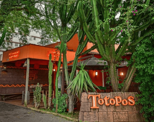 Exterior de um restaurante laranja vibrante chamado Totopos, aninhado entre grandes cactos e vegetação exuberante em um dos bairros nobres de Curitiba, com uma placa de madeira exibindo o nome do restaurante em letras grandes.