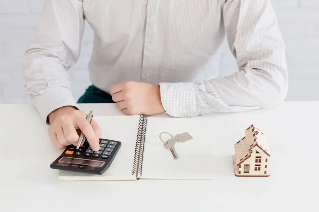 Imagem de uma pessoa de camisa clara usando uma calculadora em uma mesa com um caderno, chaves de casa e uma pequena maquete de casa