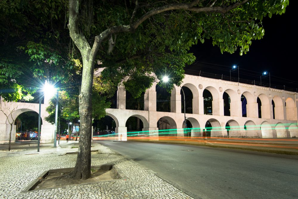 Uma cena noturna do aqueduto dos Arcos da Lapa, no Centro do Rio de Janeiro, com uma grande árvore em primeiro plano, postes de luz brilhando e rastros de luz dos veículos que passam em uma rua tranquila.