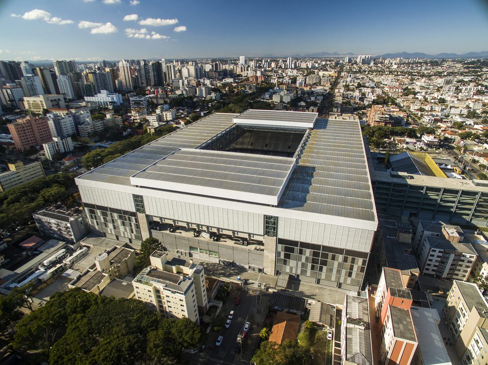 Vista aérea de um grande e moderno centro de convenções cercado por prédios altos e paisagem urbana no Morar em Curitiba, com um telhado em forma de grade e paisagem urbana se estendendo à distância sob um céu parcialmente nublado.