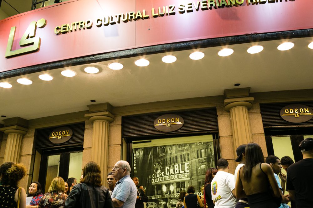 Uma multidão se aglomera em frente ao Centro Cultural Luiz Severiano Ribeiro, o Odeon, no Centro do Rio de Janeiro, sob uma marquise vermelha iluminada à noite. O prédio tem grandes janelas e portas, e as pessoas conversam em grupos.