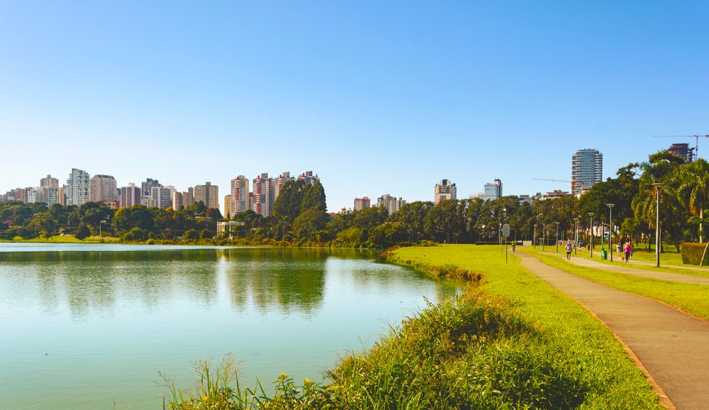 Um parque municipal com um lago e uma trilha para caminhada, cercado por grama verde e árvores, mostra por que muitos escolhem morar em Curitiba — prédios altos se erguem ao fundo sob um céu azul claro.