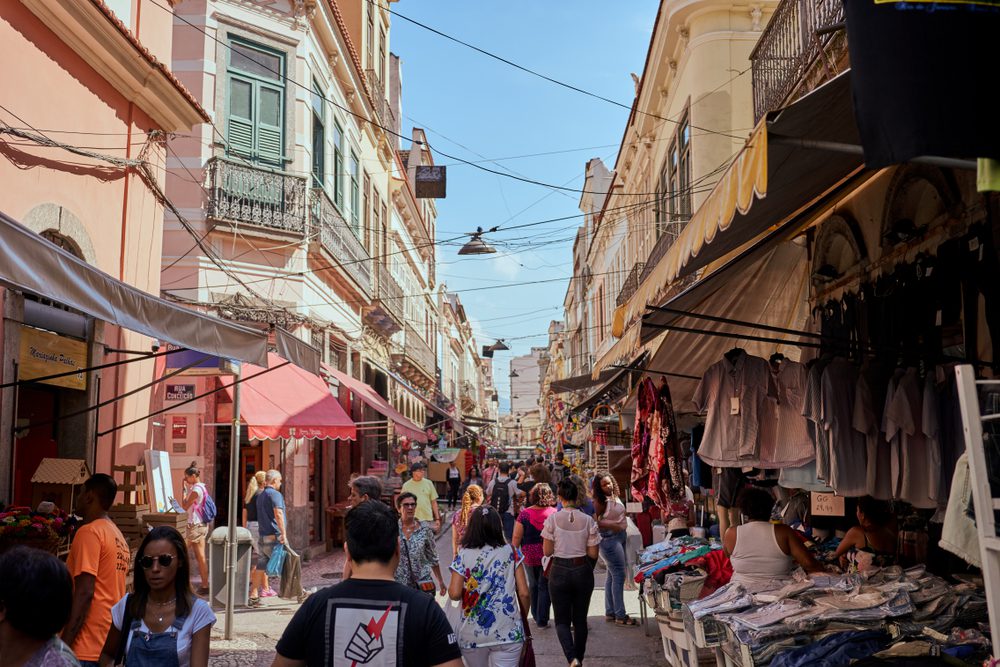 Uma movimentada rua de mercado ao ar livre no Centro do Rio de Janeiro em um dia ensolarado, com pessoas caminhando, fazendo compras e olhando diversas barracas de roupas, cercadas por coloridos edifícios históricos.