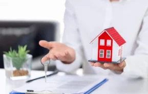 Imagem das mãos de uma pessoa segurando uma casa de madeira em tamanho miniatura e as chaves de um imóvel para ilustrar matéria sobre a averbação de imóvel