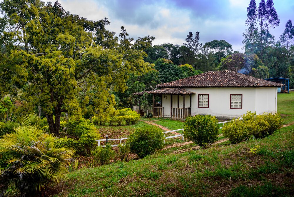 Uma casa branca com telhado de telhas fica em uma paisagem verdejante com árvores, arbustos, um pequeno jardim e um caminho que leva à varanda sob um céu nublado — perfeito para quem busca casas rústicas charmosas.