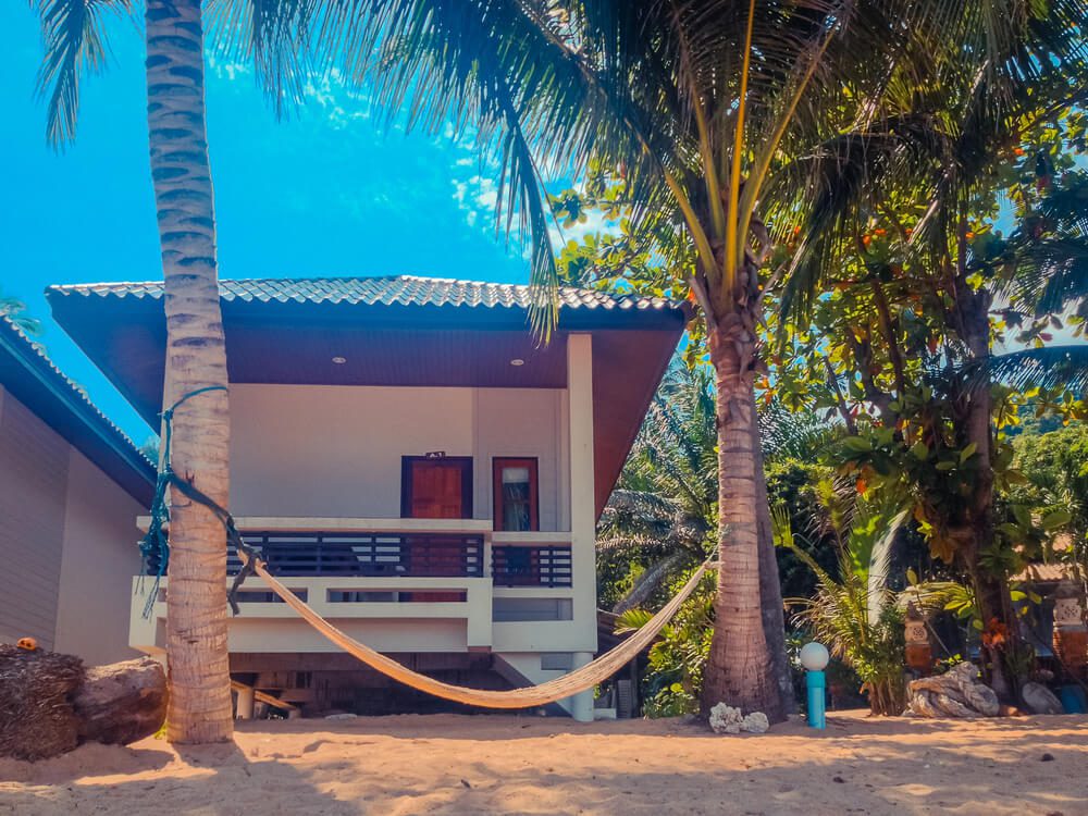 Um aconchegante bangalô de praia inspirado em casas rústicas, com telhado de telhas, varanda e rede entre duas palmeiras, situado em terreno arenoso com vegetação tropical exuberante e um céu azul brilhante.