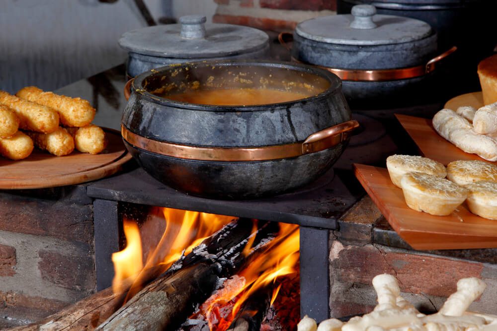 Uma panela preta com alças de cobre cozinha um ensopado em fogo de lenha em um fogão rústico, evocando o calor e o charme das casas rústicas, cercada por vários pães e acompanhamentos em tábuas de madeira.
