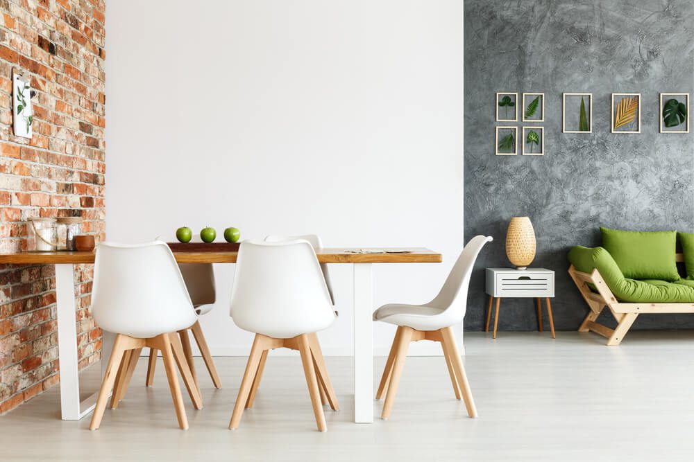 Uma sala de jantar moderna com mesa de madeira, cadeiras brancas e parede de tijolos aparentes evoca o charme das casas rústicas. Ao fundo, um sofá verde, uma mesa lateral com luminária e uma arte emoldurada com folhas decoram uma parede cinza texturizada. Maçãs verdes estão sobre a mesa.