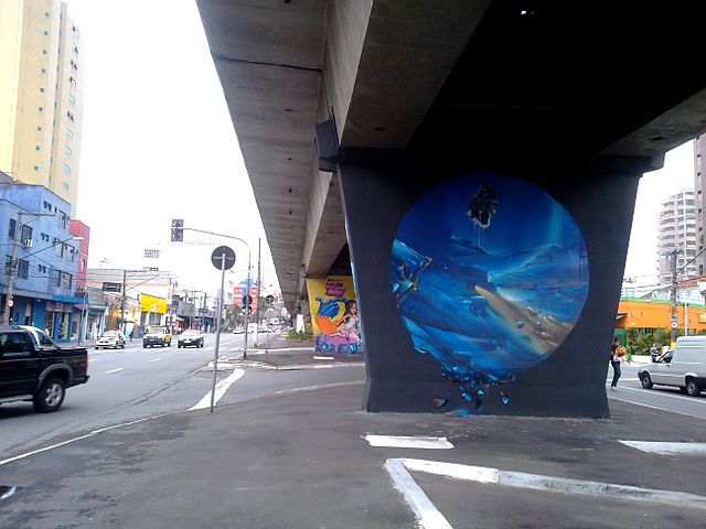640px-MAAU-SP2 Uma cena de rua mostra um viaduto com um pilar com um grande mural circular de uma cena subaquática, cercado por prédios, carros e grafite colorido em SP ao fundo.
