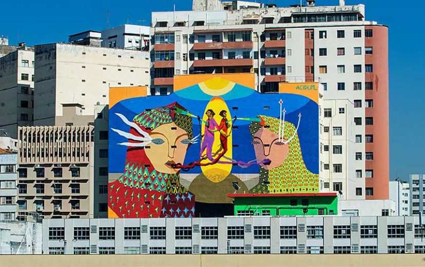 Um mural colorido em um prédio da cidade em Grafite em BH apresenta dois rostos grandes e estilizados com padrões geométricos e uma figura menor com asas entre eles. Prédios altos e modernos circundam o mural sob um céu azul claro.