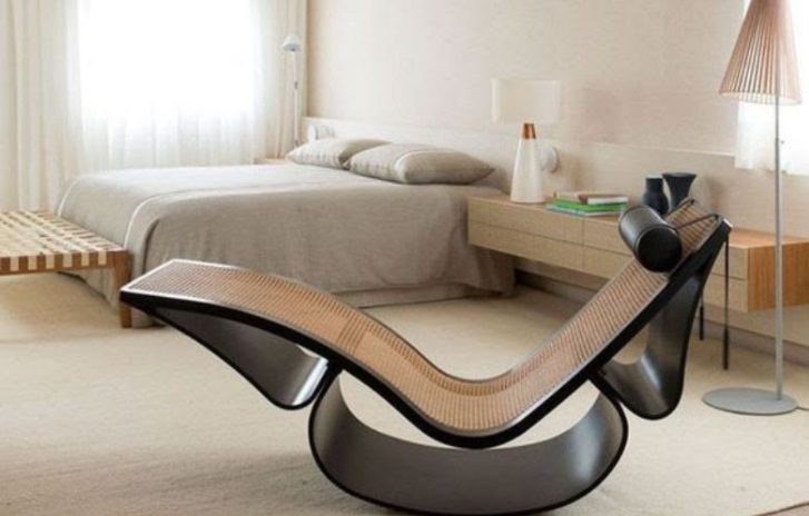 Um quarto moderno com uma cama bege e roupa de cama combinando, um criado-mudo com luminária e uma elegante chaise longue preta e de rattan, inspirada nos móveis modernistas. O quarto tem um design minimalista e aconchegante.