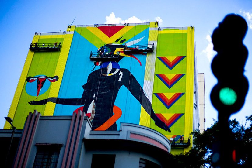 Um mural colorido cobre a lateral de um prédio alto no Grafite em BH, retratando uma figura abstrata e estilizada segurando um símbolo do útero em meio a formas geométricas e cores vibrantes. Um semáforo verde se destaca em primeiro plano.