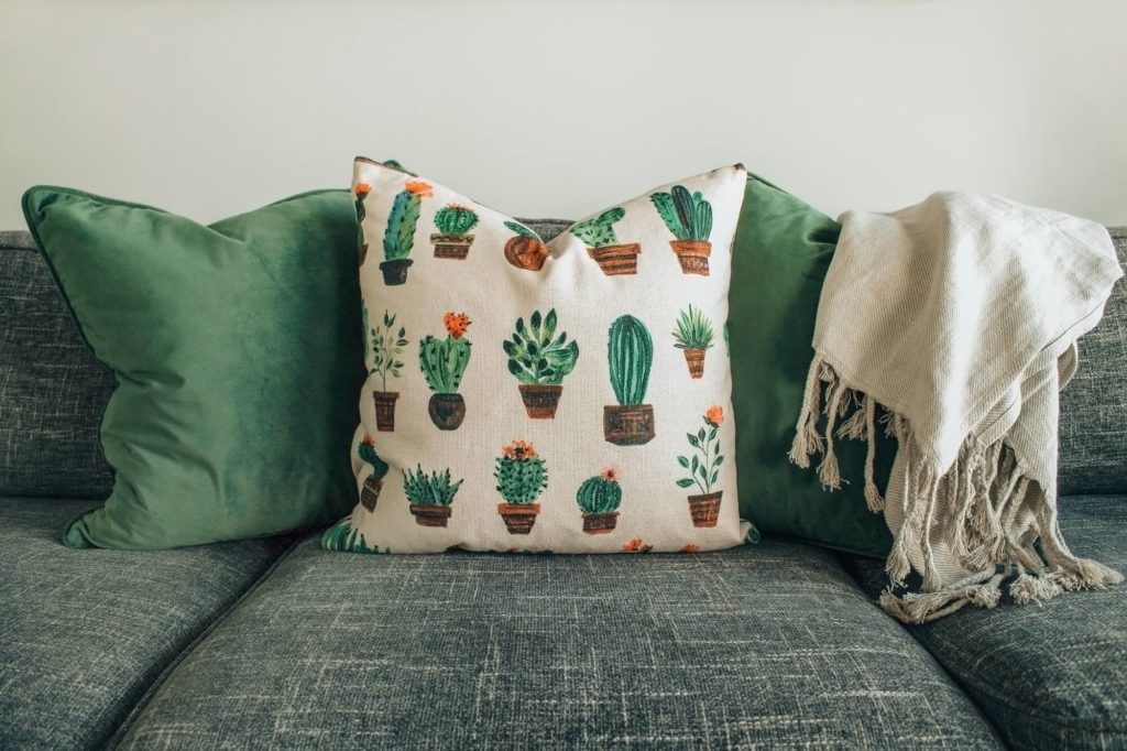 Um sofá cinza com almofadas para sofá em verde, uma almofada decorativa com ilustrações de cactos em vasos e uma manta com franjas bege claro sobre o apoio de braço.