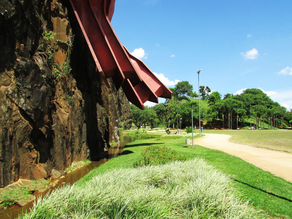 Uma escultura de metal vermelho se projeta de um penhasco rochoso ao lado de um lago e trilha em um dos Parques em Campinas, cercada por grama verde, plantas e árvores sob um céu azul claro.
