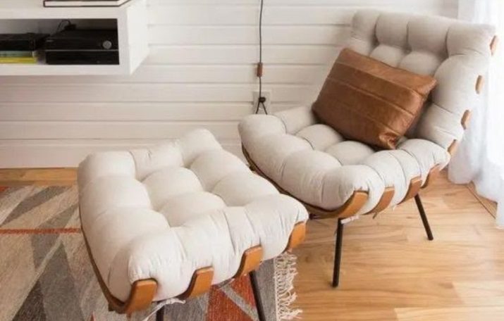 Uma poltrona aconchegante e estofada com pufe combinando em tecido bege claro e acabamento em couro marrom, inspirada em móveis modernistas, fica sobre um piso de madeira ao lado de uma janela, com uma almofada marrom e um tapete estampado por perto.