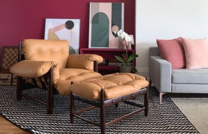 Uma sala de estar aconchegante com móveis modernistas, incluindo uma poltrona de couro bege e um pufe sobre um tapete preto e branco. Há um sofá cinza com almofadas rosa, arte moderna, uma planta e uma parede com detalhes em vermelho escuro ao fundo.