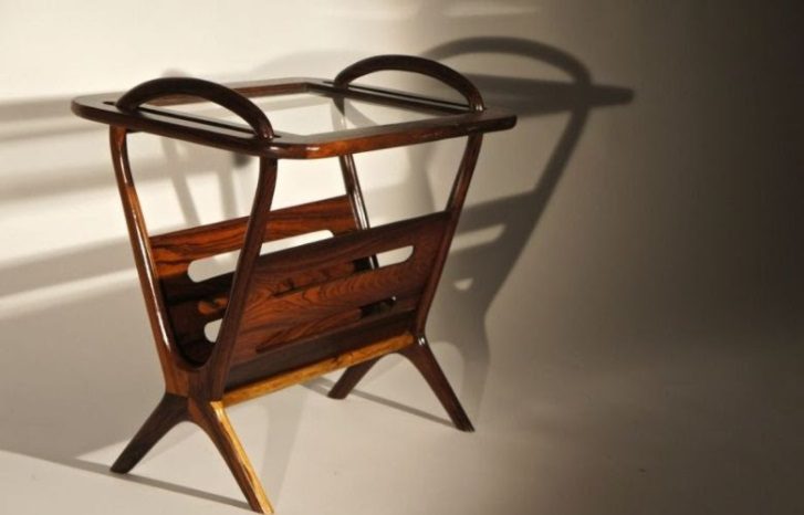 Um porta-revistas de madeira moderno de meados do século, com alças curvas e pernas anguladas, inspirado em móveis modernistas, projeta uma sombra distinta sobre um fundo claro. O porta-revistas possui tampo de vidro e acabamento em madeira escura polida.