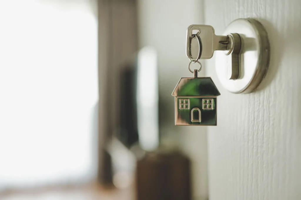 Imagem de uma chave prateada com um chaveiro em forma de casa inserida em uma fechadura de porta com um fundo desfocado sugerindo o interior de uma casa